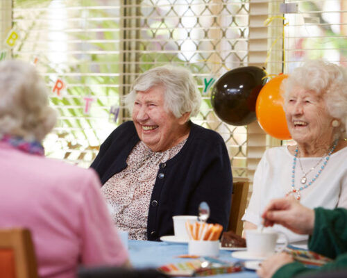 Two ladies laughing and smiling at a shared table with balloons behind.